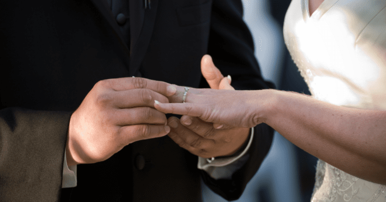 A man putting a wedding ring onto a woman wearing a wedding dress. Their faces and lower bodies are not visible.