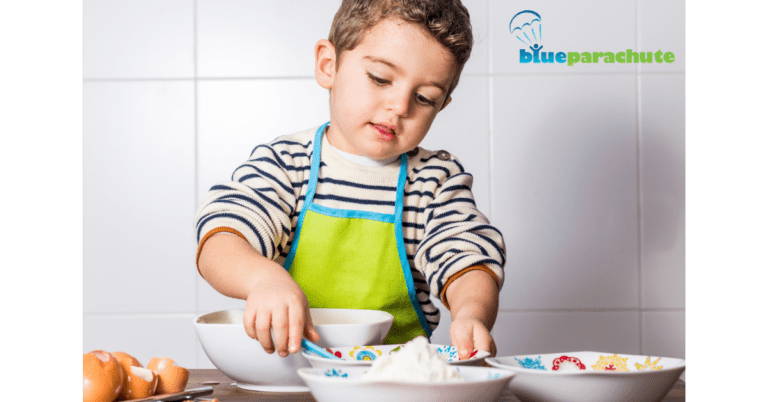 A young boy wearing a striped sweater with a green apron with blue trim worn on top us three smaller bows, one larger bowls, and some shells from brown eggs in front of him. He is grabbing one of the smaller bowls with his left hand, and about to grab a blue spoon, which is laying inside that bowl, with his right hand. This illustrates teaching how to make snacks when someone is on the autism spectrum.