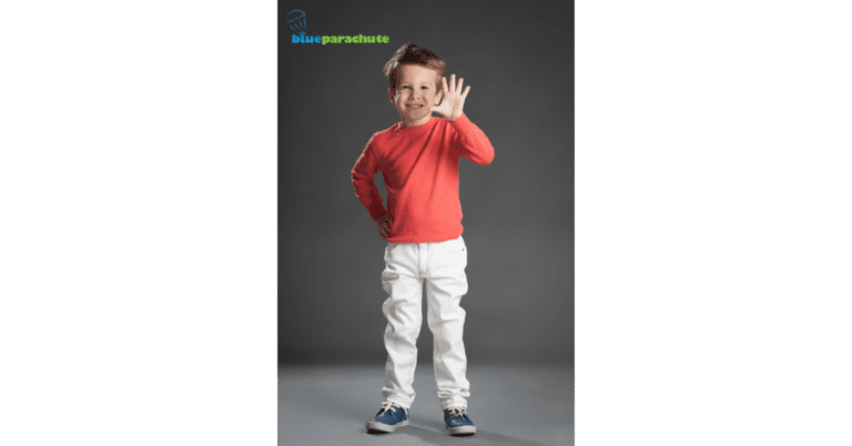 A young boy wearing a red long-sleeved shirt, white pants, and sneakers, is waving his left hand. This implies that he is being taught how to respond to hello.